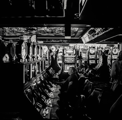 A dimly lit casino filled with rows of slot machines, some occupied by people. The machines' lights contrast with the dark surroundings, creating a vibrant yet subdued atmosphere.