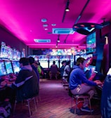 A vibrant arcade with rows of gaming machines adorned with colorful screens. Several people are immersed in playing games, seated on stools in a dimly lit room accented by an intense pink ceiling light. The atmosphere is lively and engaging.