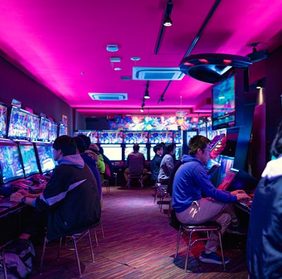 A vibrant arcade with rows of gaming machines adorned with colorful screens. Several people are immersed in playing games, seated on stools in a dimly lit room accented by an intense pink ceiling light. The atmosphere is lively and engaging.