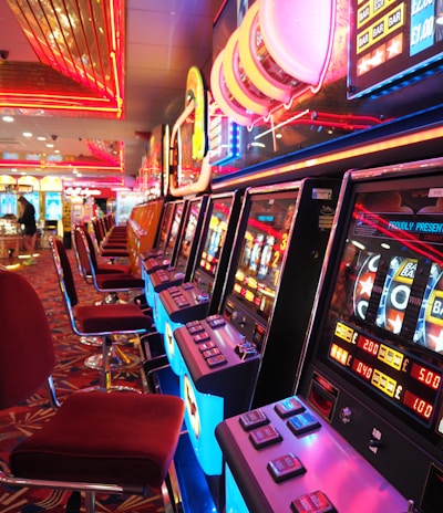 A brightly lit casino interior featuring a row of colorful slot machines. The neon lights create a vibrant atmosphere, with patterned carpeting adding to the decor. Stools are aligned neatly in front of each machine.