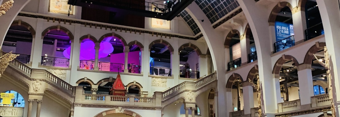 The interior of a large, ornately designed building with arched ceilings and intricate architectural details. The space features gaming machines lined up along the floor, under a high ceiling with a grid pattern. Lighting in purple and white tones enhances the ambiance.