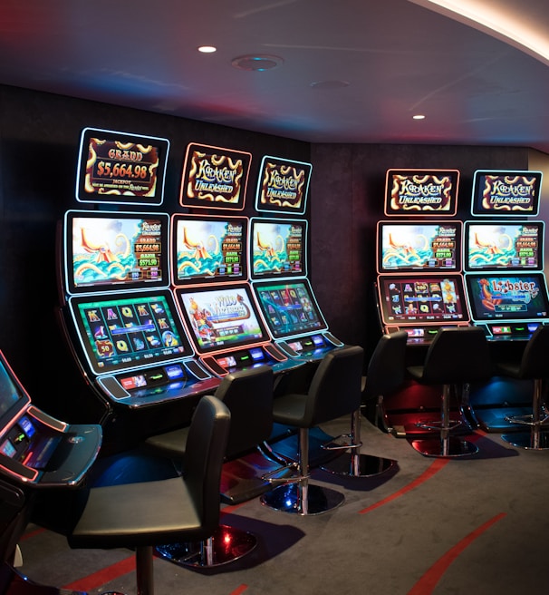A row of brightly lit slot machines in a casino setting, each with colorful displays and various game themes. The machines are lined up against a dark wall and are unoccupied, with cushioned chairs placed in front of them. The area is dimly lit except for the glow from the screens, creating a vibrant and engaging atmosphere.