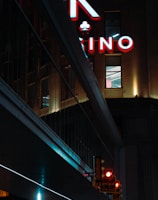 A night scene featuring a building with illuminated neon signage displaying 'CASINO'. The environment is urban with reflections on glass windows and glowing street traffic lights below. The atmosphere is dimly lit, with artificial lighting casting vibrant colors.