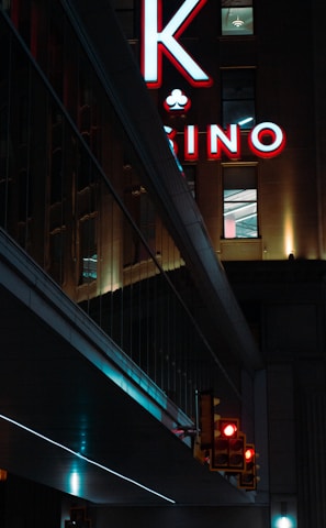 A night scene featuring a building with illuminated neon signage displaying 'CASINO'. The environment is urban with reflections on glass windows and glowing street traffic lights below. The atmosphere is dimly lit, with artificial lighting casting vibrant colors.